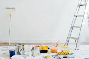 Painting tools including rollers, brushes, paint cans, and a ladder arranged on the floor of a room being painted with a white wall in the background