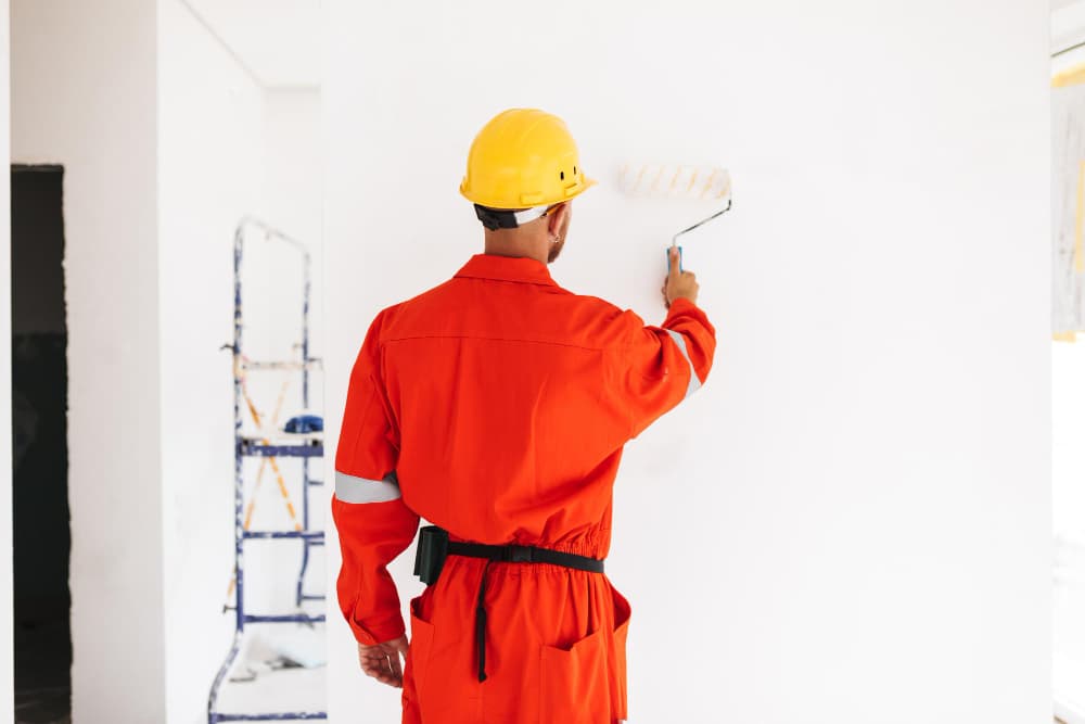 Professional painter in a red uniform and yellow hard hat applying paint to an interior wall using a roller in a bright, unfinished room
