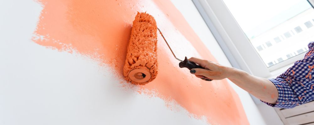 A close-up of a person using a paint roller to apply bright orange paint to a white interior wall