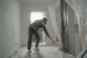 A professional painter in protective gear using a high-efficiency spray gun in a fully masked room, illustrating the specialized labor and equipment that factor into the cost to paint a house interior.