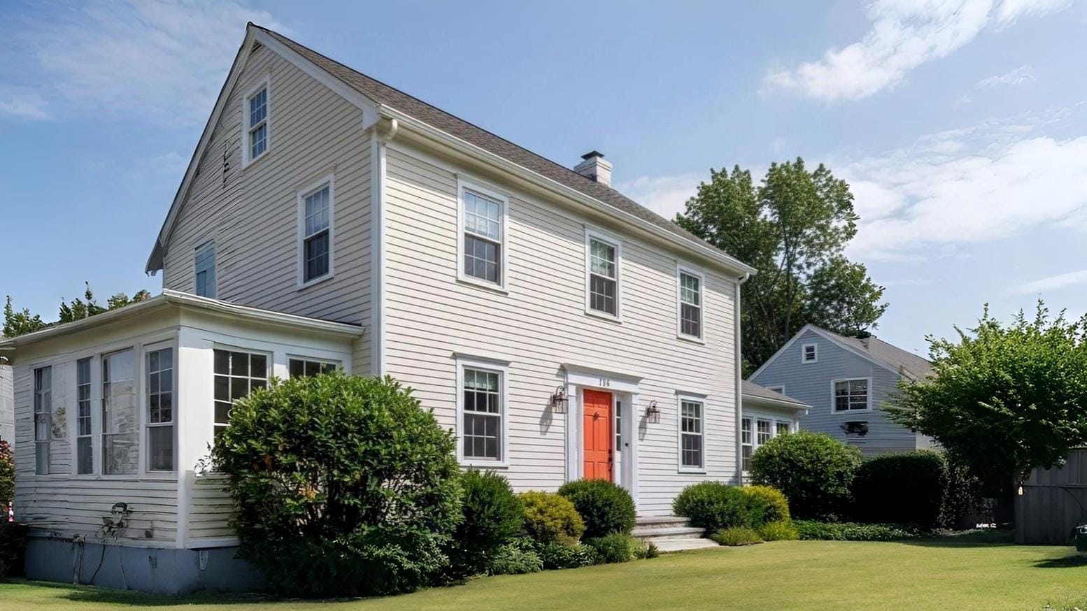 A side-angled view of a two-story cream-colored house featuring a vibrant orange front door, white trim, and dark grey roofing shingles. The home is surrounded by lush green grass and a paved stone walkway.