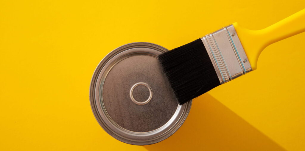 A top-down view of a closed paint can and a yellow-handled brush on a vibrant yellow surface, illustrating how the best paint finishes can transform a space with bold, saturated color.