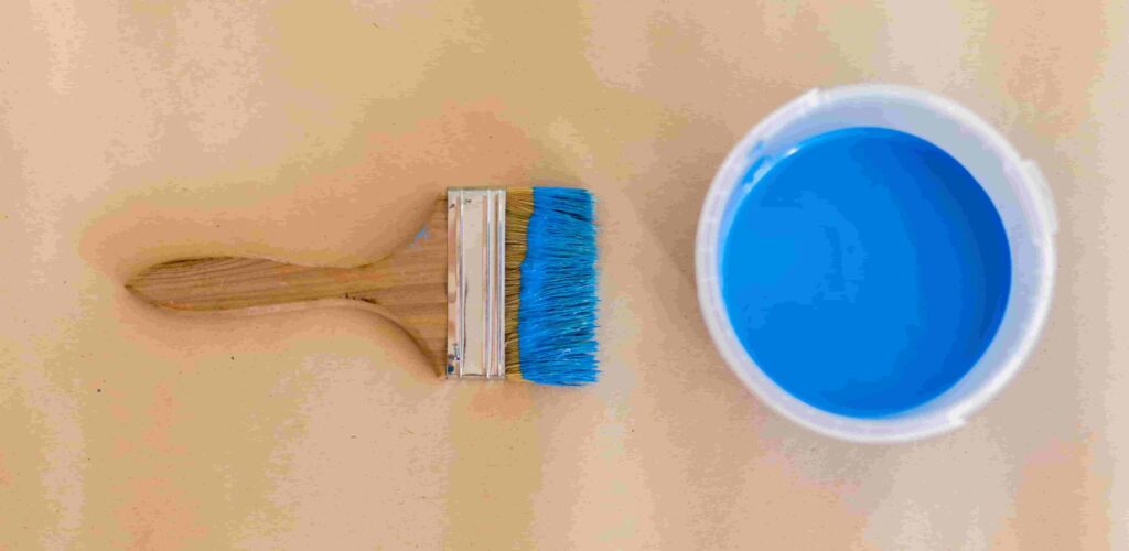 A flat-lay view of a wooden paint brush and a bucket of vibrant blue interior paint, perfect for demonstrating how the best paint finishes apply smoothly to residential surfaces.